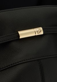 Section d'un sac en cuir noir présentant une fermeture à bouton pression dorée avec un logo. Texture lisse et détails de coutures contrastantes soulignent son design.