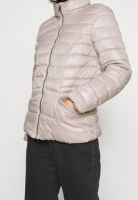 Jasno beżowa kurtka puchowa z wysokim kołnierzem, zamkiem błyskawicznym i pikowanym wzorem. Posiada kieszenie boczne oraz gładką, błyszczącą fakturę.
