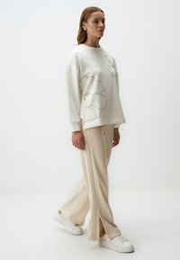 Sweatshirt cream avec un motif floral en contour, associé à un pantalon beige avec des fentes latérales et des baskets blanches. Style minimaliste, coupe décontractée.