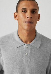 Ljusgrå polo skjorta gjord av texturerat tyg, med en spridd krage och två vita knappar vid knäppningen. Ingen mönster eller dekorationer.