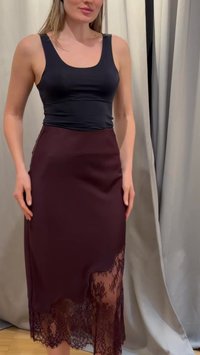 Femme portant un haut noir sans manches et une jupe bordeaux foncé avec des détails en dentelle à l’ourlet, debout devant un fond de rideau neutre.