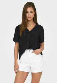 Svart blus med textur, korta ärmar och V-hals, matchad med vita denimshorts. Blusen har ett diskret prickigt mönster.