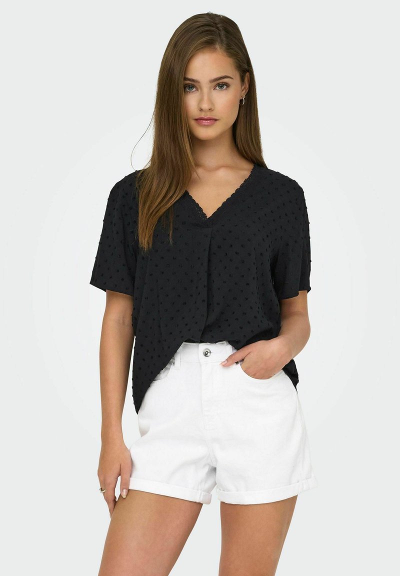 Svart blus med textur, korta ärmar och V-hals, matchad med vita denimshorts. Blusen har ett diskret prickigt mönster.