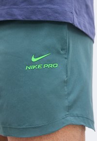 Κοντινό πλάνο σε τιρκουάζ σορτς Nike Pro με φωτεινό πράσινο λογότυπο Nike και κείμενο στο μηρό ενός ατόμου που φοράει μπλε πουκάμισο.