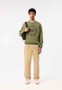 Felpa verde oliva con logo circolare, collo a giro in crema, abbinata a pantaloni beige e sneakers chiare. Borsa nera appesa a una spalla.