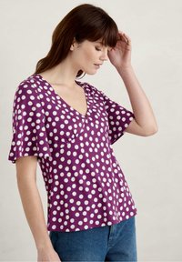 Fioletowy krótki rękaw bluzka z dekoltem w serek, zdobiona białymi kropkami, z rozkloszowanymi rękawami i luźnym krojem. Miękka tkanina o gładkiej fakturze.