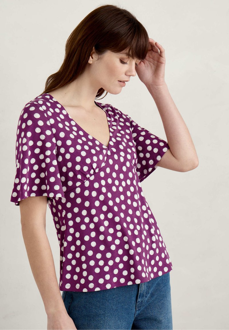 Fioletowy krótki rękaw bluzka z dekoltem w serek, zdobiona białymi kropkami, z rozkloszowanymi rękawami i luźnym krojem. Miękka tkanina o gładkiej fakturze.