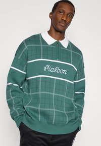 Πράσινο πλεκτό πουλόβερ με λευκό γιακά, με σχέδιο πλέγματος και κεντημένο λογότυπο "Malbon". Λεπτομέρεια φερμουάρ μπροστά και μαλακή υφή.