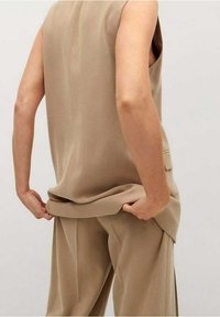 Beiges ärmelloses Oberteil mit lockerer Passform, das über dunkelbeige weit geschnittenen Hosen liegt. Weiche, glatte Textur mit lässigem Design.