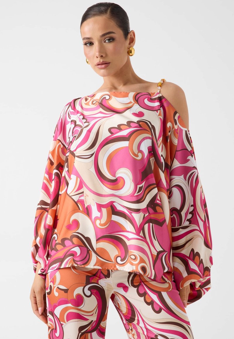 Femme portant une tenue à motif abstrait tourbillonnant rose vif, orange et blanc avec une découpe asymétrique à une épaule et des boucles d'oreilles anneaux dorés.