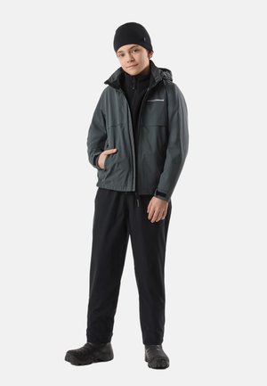 Jeune personne portant un bonnet noir, une veste à capuche grise, un pantalon noir et des chaussures noires, debout avec une main dans la poche de la veste.