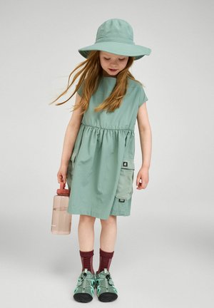 Młoda dziewczyna w zielonej sukience i kapeluszu z szerokim rondem, trzymająca butelkę z wodą, ubrana w bordowe skarpetki i zielone sandały, patrząca w dół.