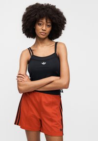 Μαύρο αθλητικό τοπ με λεπτές τιράντες και λευκό λογότυπο στο κέντρο, σε συνδυασμό με πορτοκαλί σορτς με μαύρες πλευρικές ρίγες και λογότυπο.