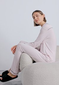 Set de pijama roz deschis, cu mâneci lungi și pantaloni, având detalii din satin negru. Modelul stă pe un scaun curbat, tapițat bej. Papuci negri.