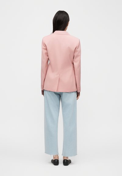 Γυναίκα με μακριά σκούρα μαλλιά που φοράει ροζ σακάκι, ανοιχτό μπλε τζιν παντελόνι με φαρδιά πόδια και ξεφτισμένες άκρες, και μαύρα παπούτσια, στέκεται γυρισμένη προς τα πίσω.