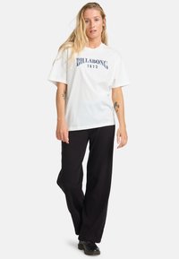 Hvid bomulds t-shirt med blå "Billabong 1973" grafik, som er parret med sorte vide bukser og båret med sorte sko.
