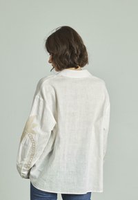 Camicia bianca in lino a maniche lunghe con dettagli in ricamo dorato sulla manica sinistra. Tessuto morbido, vestibilità rilassata e polsini con bottone.