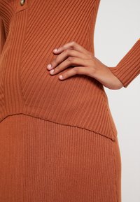 Hedvábné, pletené šaty v rezavé barvě s přiléhavým střihem, kulatým výstřihem a dlouhými rukávy, zvýrazněné mírně třepeným lemem.