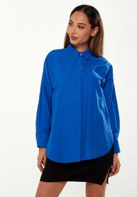 Blue button-up shirt with a collar, long sleeves, and a relaxed fit; fabric appears smooth and lightweight; paired with a black mini skirt.