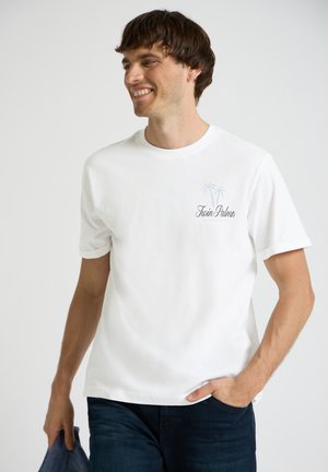 Smilende mand iført en hvid "Twin Palms" T-shirt, blå jeans, der holder en mørkeblå jakke i den ene hånd, og kigger mod venstre.