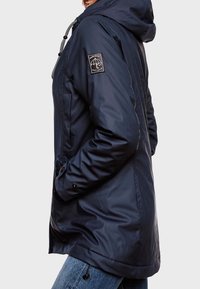 Chaqueta impermeable azul marino con capucha, que presenta un parche decorativo con logo, detalles de cordones y bolsillos laterales, hecha de un material sintético suave.