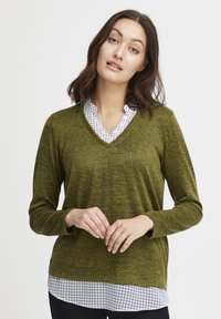 Zielona oliwkowa sweter w serek z długimi rękawami, o strukturalnym plecionym materiale. Nałożony na białą koszulę w kratę z kołnierzykiem.