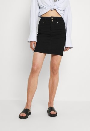 Kvinde iført nederdel i sort højhalset denim, hvid langærmet skjorte bundet ved taljen og sorte sandaler med åbne tæer.