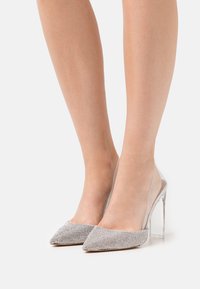 Transparent klackpumps med en silverglittrande ovandel, spetsig tådesign och klar klack. Slät yta med metalliska accenter.