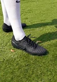 Sorte Nike fodboldstøvler med et struktureret design, der har en glat overdel, snørebånd og en lav profil sål, båret med hvide knæstrømper.
