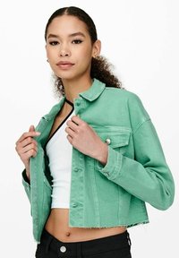 Giacca di jeans verde menta con design corto, tasche sul petto a doppio, e bottoni argentati, esposta parzialmente aperta sopra una maglietta bianca.