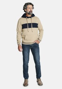 Sudadera con capucha color beige, con una franja azul marino en el pecho, bolsillo tipo canguro, combinada con jeans oscuros y calzado beige.