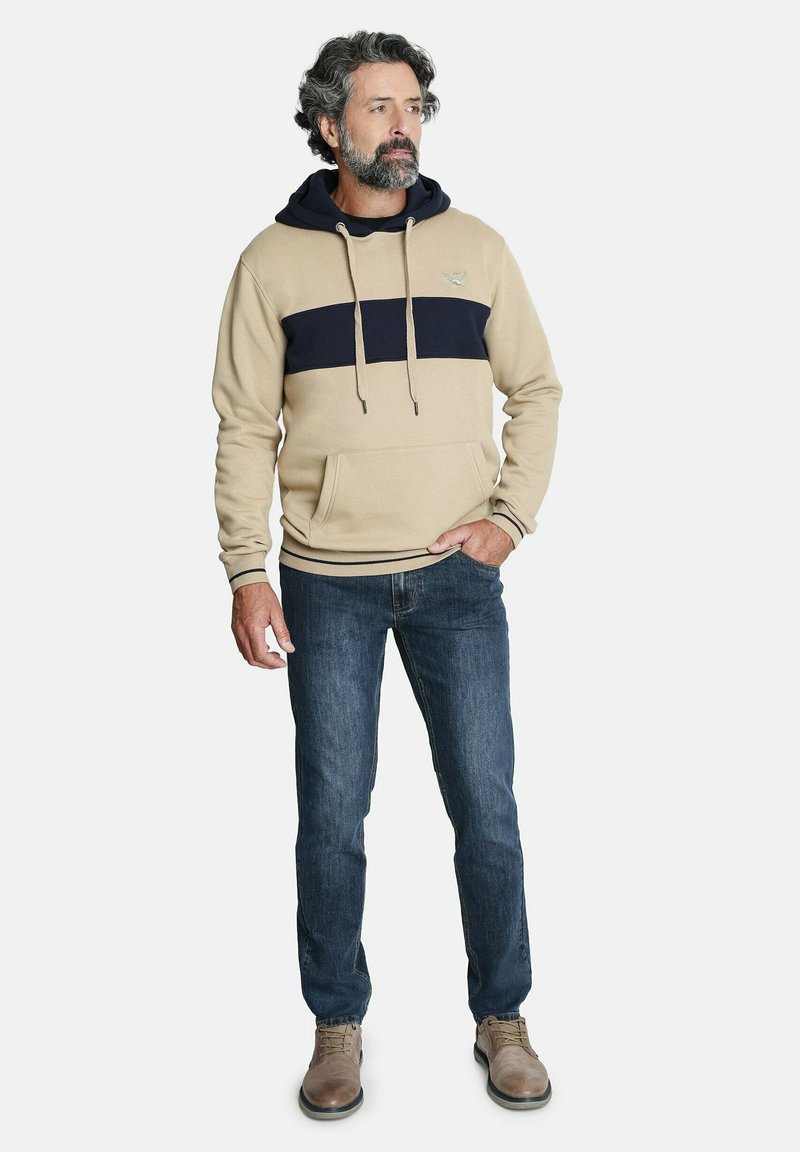 Sudadera con capucha color beige, con una franja azul marino en el pecho, bolsillo tipo canguro, combinada con jeans oscuros y calzado beige.