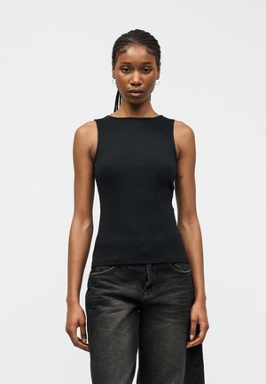 Woman wearing a sleeveless black ribbed top and faded black jeans, standing with arms relaxed against a plain white background.