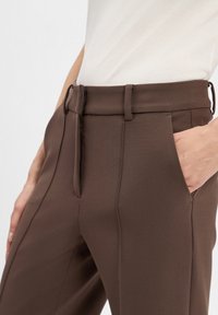 Braune maßgeschneiderte Hosen mit glatter Textur, Gürtelschlaufen und seitlichen Taschen sowie einem Reißverschluss vorne.