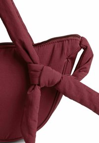 Borsa in tessuto bordeaux con manico annodato, dotata di una chiusura con zip e una superficie liscia. La forma è arrotondata con dettagli di cucitura sottili.