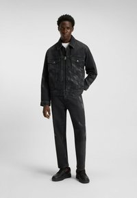 Young man standing, wearing black washed denim jacket and matching jeans with subtle diagonal patterns, black sneakers, against plain background.