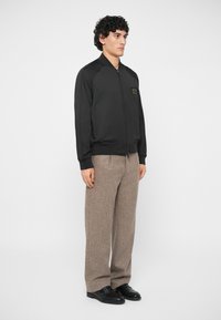 Άνδρας που στέκεται σε μαύρο φούτερ με φερμουάρ και λογότυπο, καφέ παντελόνι σε φαρδιά γραμμή και μαύρα κοστούμια παπούτσια, μπροστά σε λευκό φόντο.