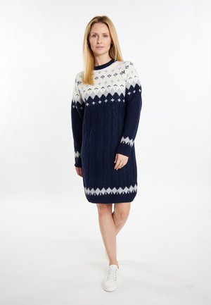 Vrouw met lang blond haar die een marineblauwe en witte gebreide sweaterjurk met patroon en witte sneakers draagt, staand tegen een witte achtergrond.
