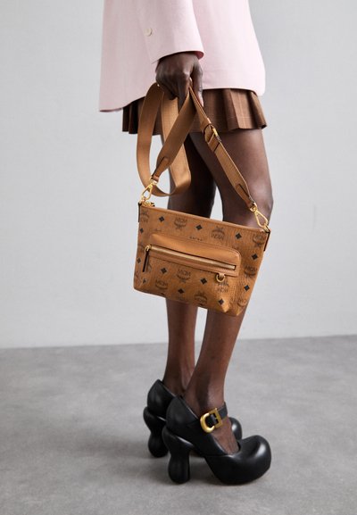 Läder crossbody-väska i tan med präglat logomönster, zip-ficka och detaljer i guldmetall. Axelhänget är justerbart och platt.