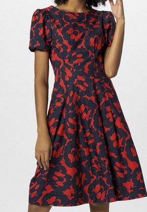 Femme portant une robe bleu marine longueur genou avec des manches ballon et des motifs floraux abstraits rouges audacieux, posant avec une main près de son visage.