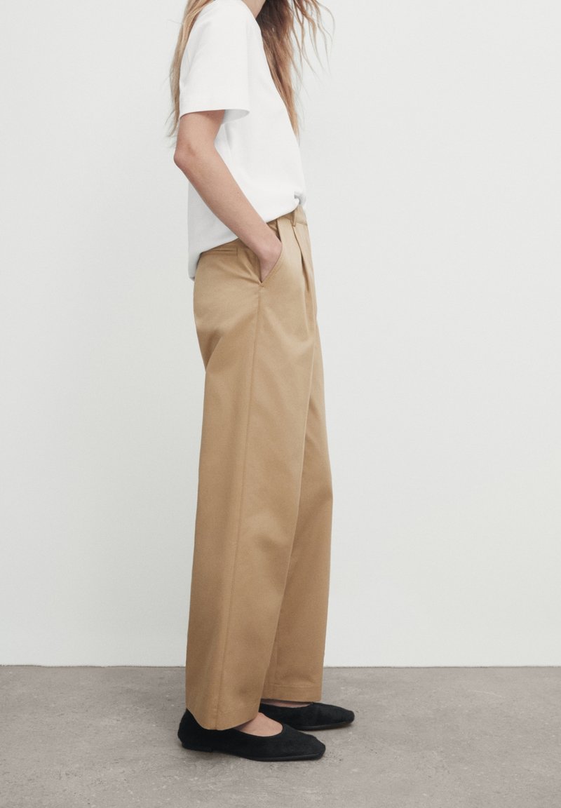 Personne portant un pantalon beige à jambes larges, un haut blanc à manches courtes, des ballerines noires, debout les mains dans les poches contre un mur blanc.