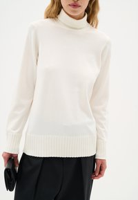 Maglione bianco a collo alto con polsini e orlo a coste, realizzato in tessuto morbido. Abbinato a pantaloni neri e tiene una borsa a mano nera.