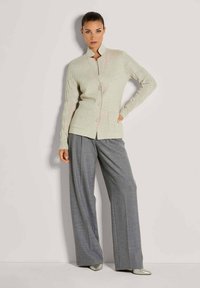 Eine Frau steht mit der Hand in der Hüfte und trägt eine beigen, knopfbeehrte Strickjacke mit Kabelstrickärmeln sowie lockere graue, weite Hosen und spitze Schuhe.