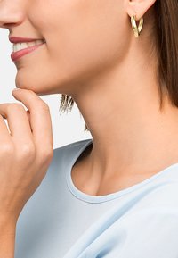 Gold hoop earrings with a twisted design, featuring a glossy finish. The model has short brown hair and wears a light blue top.