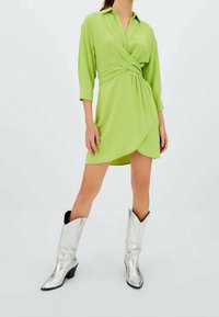 Vestito avvolgente verde a maniche lunghe in tessuto leggero, con colletto a camicia e orlo asimmetrico, abbinato a stivali da cowboy argentati.