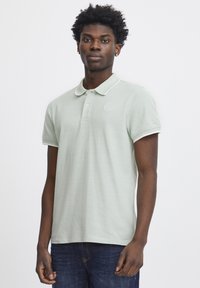 Polo shirt verde chiaro con tessuto texturizzato, maniche corte e colletto con bordo bianco. Presenta un logo nella zona del petto.