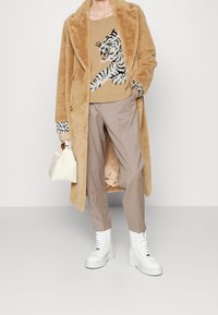 Manteau en fausse fourrure beige, pull graphique tigre beige, pantalon marron clair, bottes blanches épaisses à lacets, et petit sac à main crème.