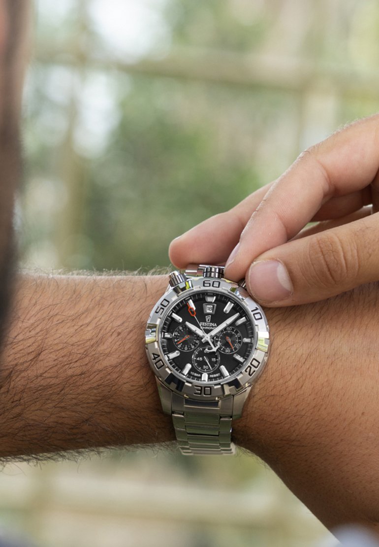 Person, die eine silberne Festina-Chronographenuhr mit schwarzem Zifferblatt am Handgelenk gegen einen verschwommenen Außenhintergrund einstellt.
