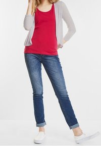 Kvinna som bär en röd linne, ljusgrå öppen cardigan, uppvikta blå jeans och vita slip-on skor, stående med ett ben korsat framåt.