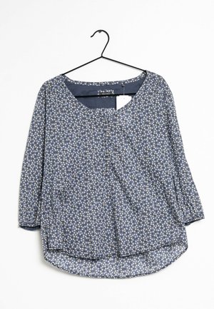 Blauwe blouse met witte en gele stippen, driekwart mouwen, knoopsluiting aan de hals, hangend aan een zwarte kledinghanger tegen een witte achtergrond.